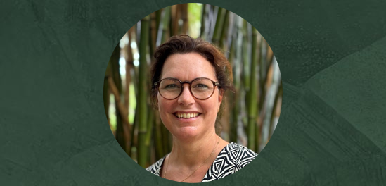 Portretfoto van Caroline Bardoel, een vrouw met kort bruin haar en een bril, lachend, gekleed in een zwart-wit patroon blouse, met een achtergrond van bamboe.